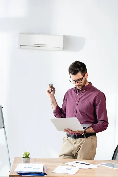 年轻商人手持笔记本电脑 用遥控器打开空调机 — 图库照片