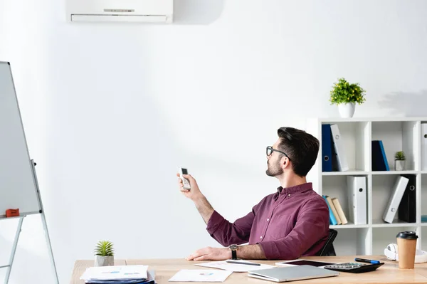 Joven Empresario Sentado Lugar Trabajo Encendiendo Aire Acondicionado Con Mando — Foto de Stock