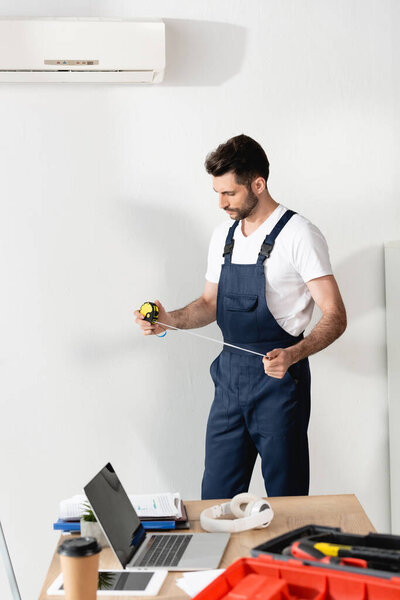 ремонтник держит измерительную ленту, стоя возле офисного стола под кондиционером
