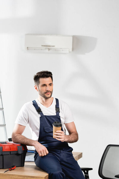 Beautiful repairman holding coffee to go while sitting at desk near toolbox under air conditioner on wall

