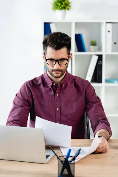 Seriöser Geschäftsmann Schaut Sich Dokumente Während Neben Laptop Sitzt — Stockfoto