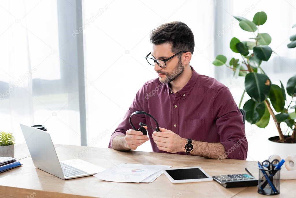 young, serious businessman holding headset while looking at laptop during video call