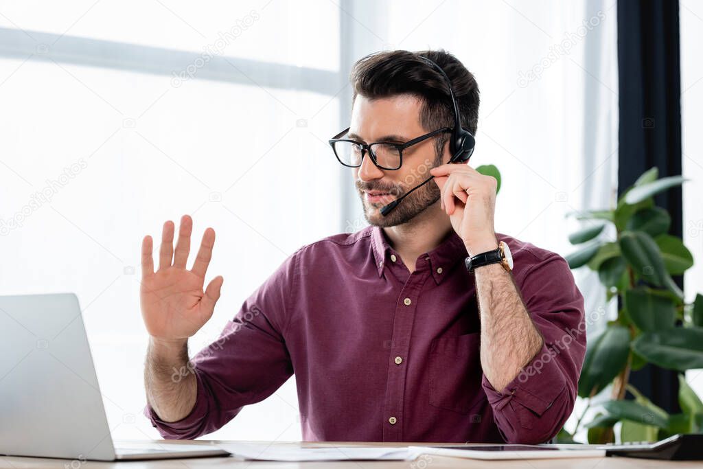smiling businessman in headset waving hand during video conference on laptop