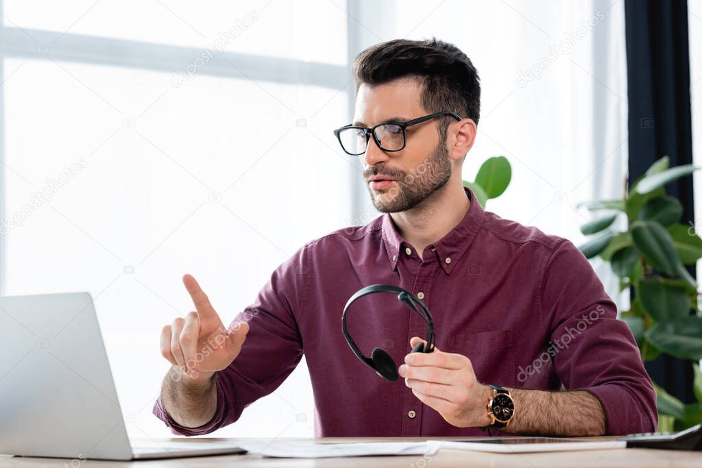 serious businessman holding headset and showing idea sign during video call on laptop