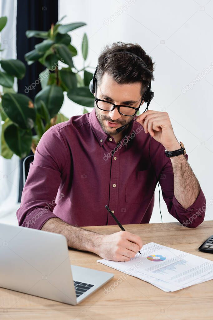 attentive businessman in headset writing on paper during online meeting on laptop