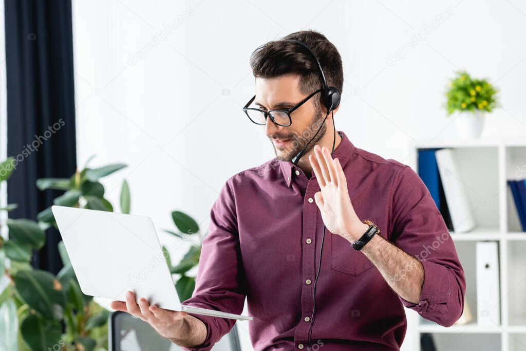 smiling businessman in headset showing hello gesture during video call on laptop