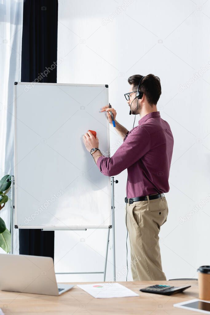 young businessman in headset writing on flipchart near desk with laptop