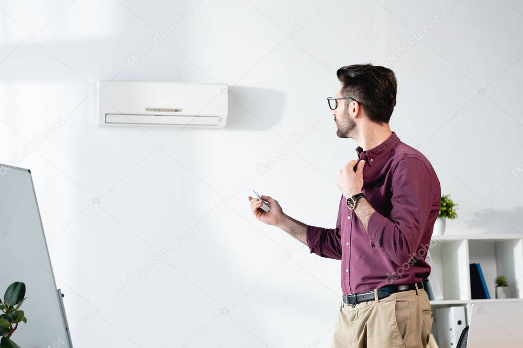 young businessman suffering from heat, touching shirt and switching on air conditioner with remote controller