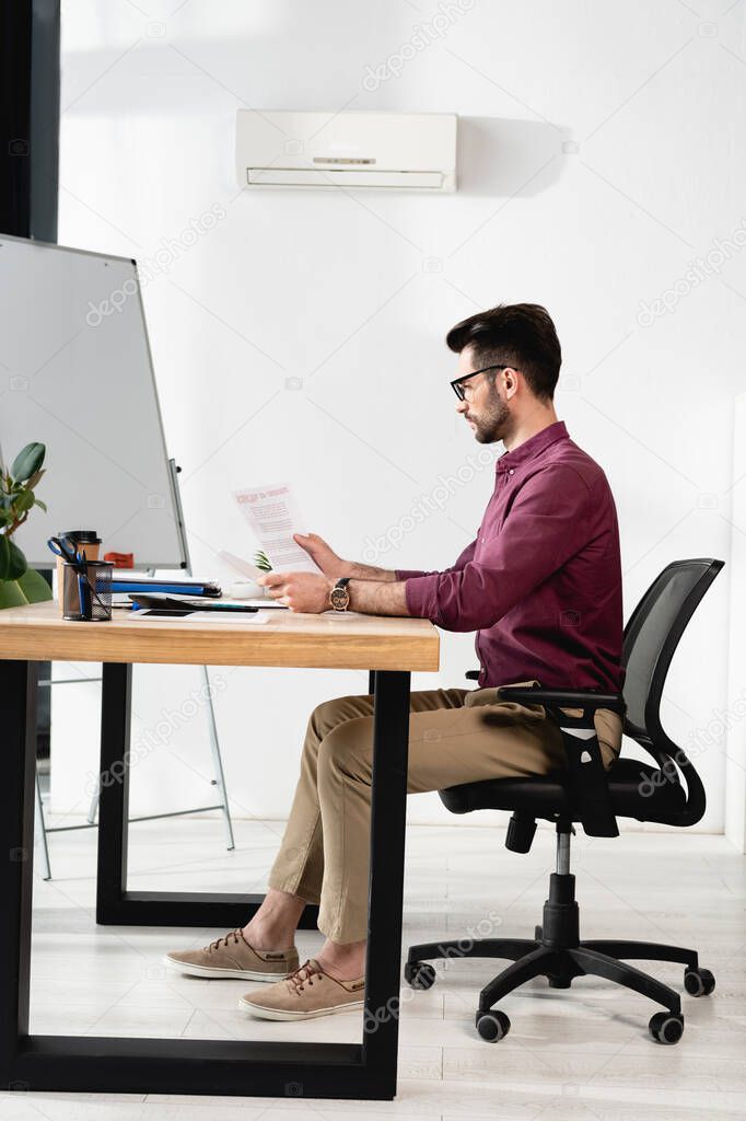 side view of businessman looking at papers while sitting at workplace near air conditioner 