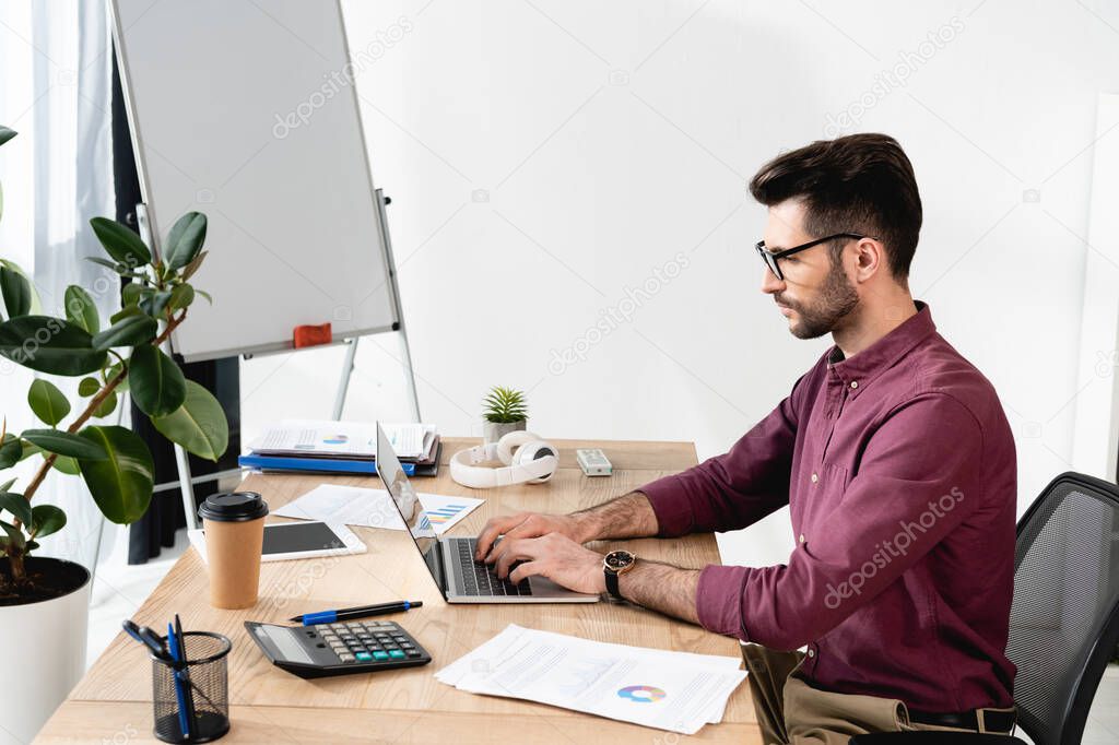 serious businessman working on laptop near documents, calculator and coffee to go