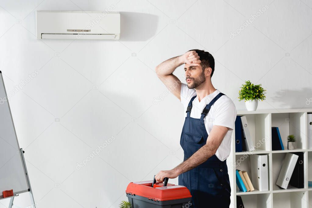 workman touching forehead while standing with toolbox near broken air conditioner and suffering from heat
