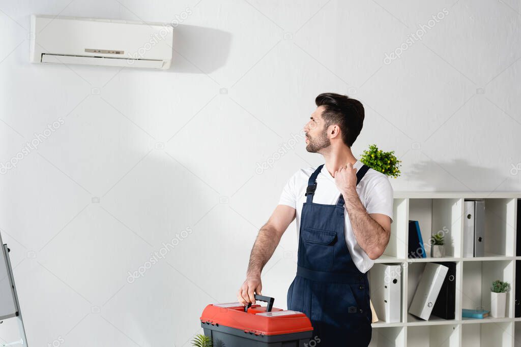 young repairman looking at air conditioner on wall while holding toolbox