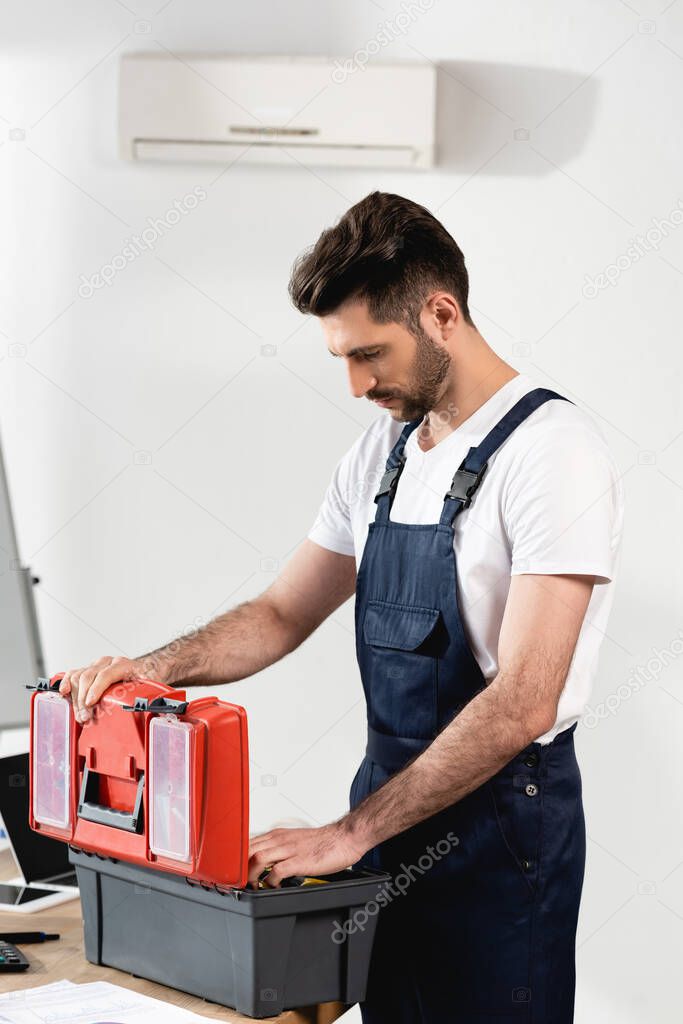 repairman in uniform opening toolbox near broken air conditioner on wall