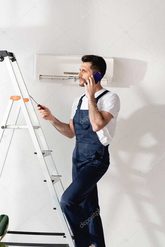 workman talking on smartphone and holding screwdriver on stepladder near air conditioner