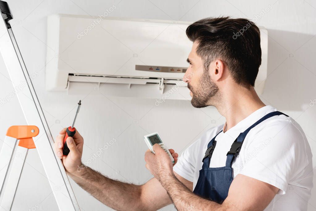 young repairman looking at air conditioner while holding remote controller and screwdriver