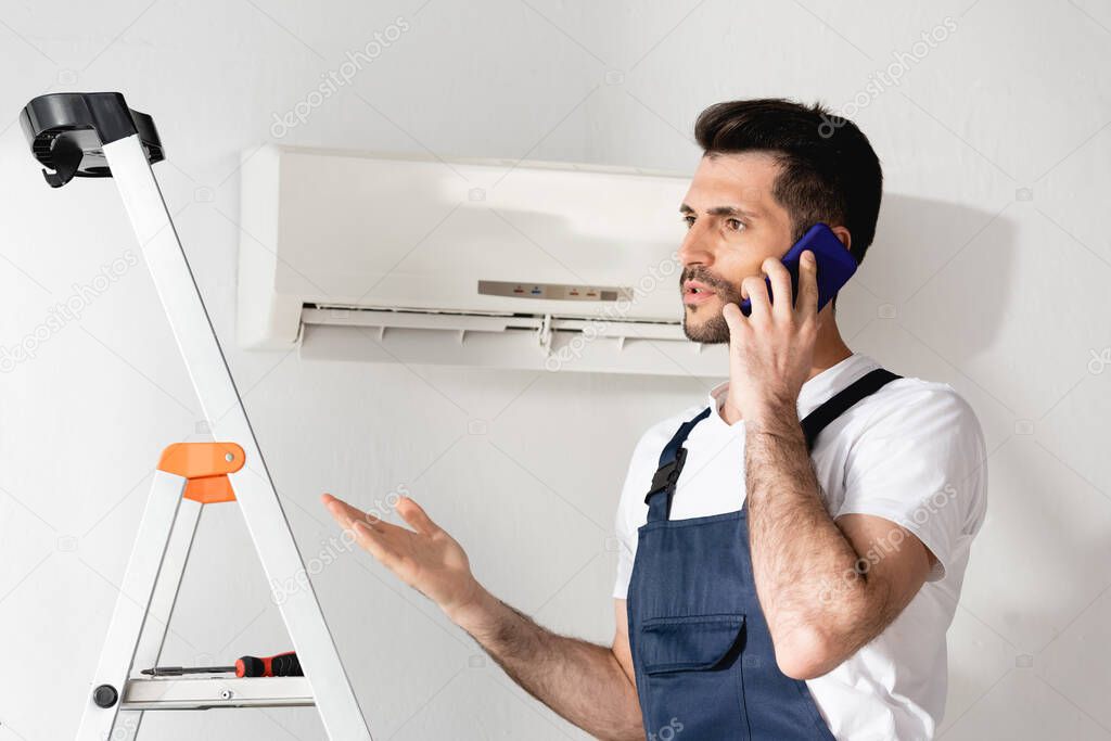 repairman standing with open arm and talking on smartphone near stepladder and air conditioner