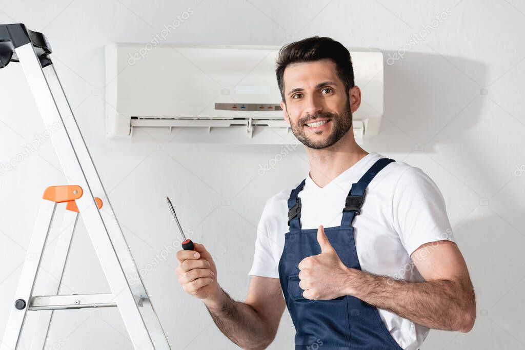 happy repairman showing thumb up and holding screwdriver while standing near stepladder and air conditioner