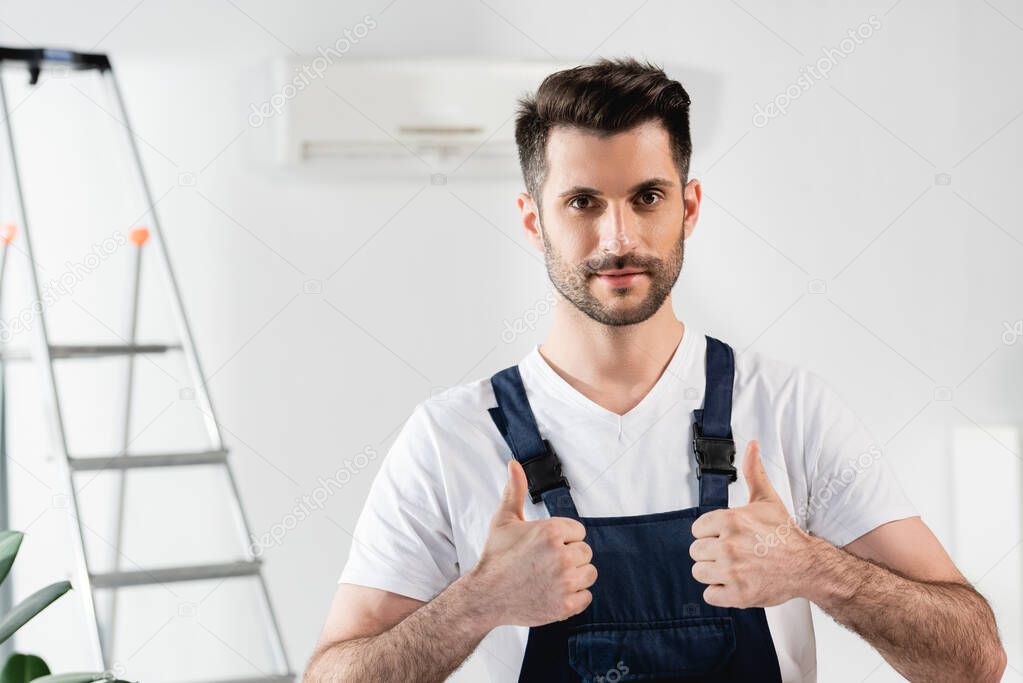 handsome repairman showing thumbs up while standing near stepladder and air conditioner on wall