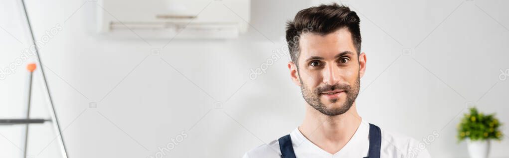 horizontal image of handsome, smiling repairman looking at camera near air conditioner on wall