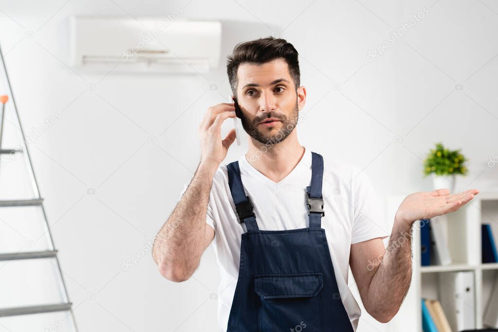 confused repairman talking on smartphone and showing shrug gesture near air conditioner on wall