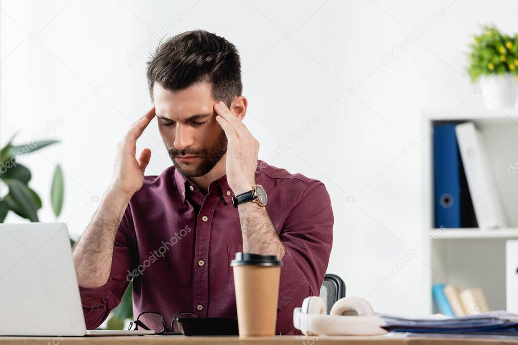 exhausted businessman with closed eyes touching head while sitting near laptop and coffee to go