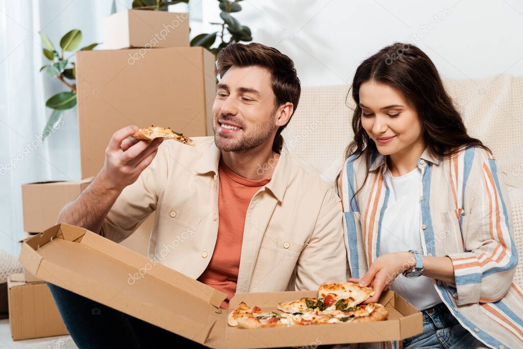 Smiling man holding piece of pizza near beautiful girlfriend and cardboard boxes at home