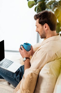 Side view of man holding coffee cup and looking at laptop on couch  clipart