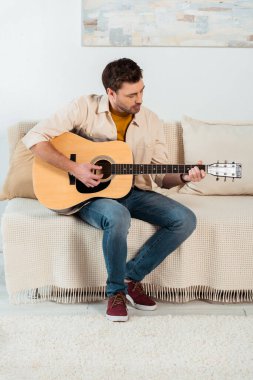 Genç adam oturma odasındaki kanepede akustik gitar çalıyor. 