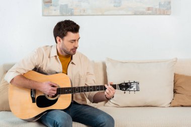 Oturma odasındaki kanepede akustik gitar çalan yakışıklı adam.