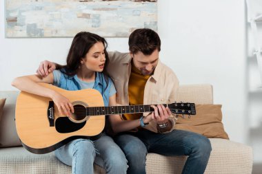 Erkek, kız arkadaşına oturma odasında akustik gitar çalmayı öğretiyor.