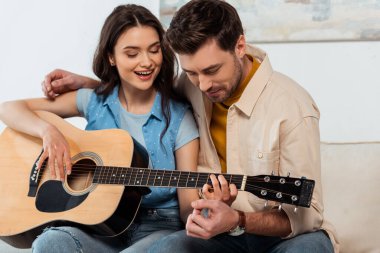 Adam akustik gitar çalarken gülümseyen kız arkadaşını kucaklıyor.