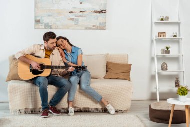 Evdeki kanepede gülümseyen kadının yanında akustik gitar çalan yakışıklı bir adam. 