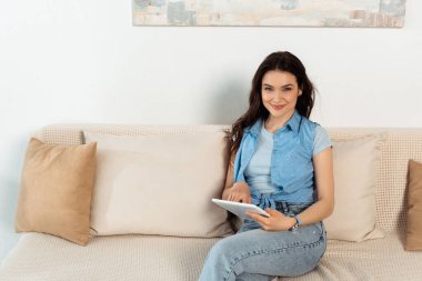 Kanepede dijital tablet kullanırken kameraya gülümseyen genç bir kadın. 