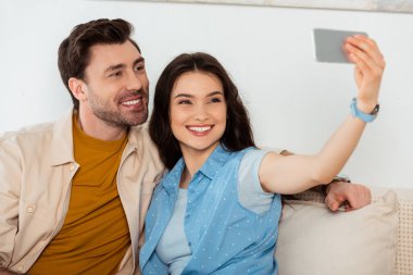 Evde gülümseyen bir erkek arkadaşın yanında akıllı telefondan selfie çeken bir kadının seçmeli odağı. 