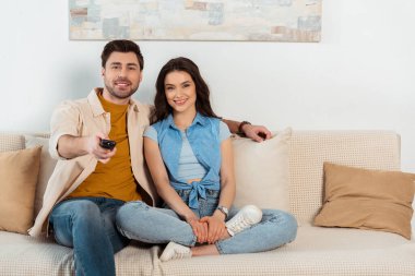 Gülümseyen çift evde televizyon izlerken kameraya bakıyor. 