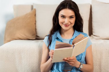 Young woman smiling at camera while reading book in living room clipart