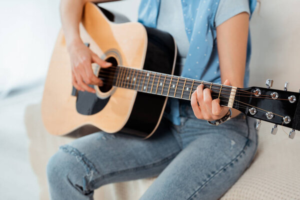 Cropped view of young woman performing on acoustic guitar on sofa 