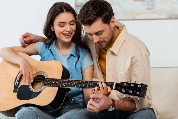 Adam akustik gitar çalarken gülümseyen kız arkadaşını kucaklıyor.