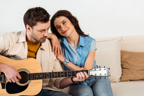 Smiling woman hugging and looking at boyfriend performing on acoustic guitar at home 