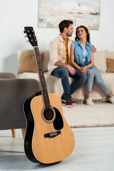 Selective focus of acoustic guitar on floor near armchair and man embracing girlfriend at home 