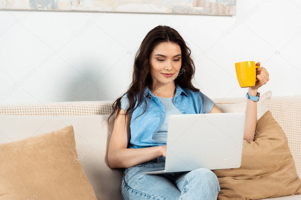 Attractive freelancer holding coffee cup and using laptop in living room