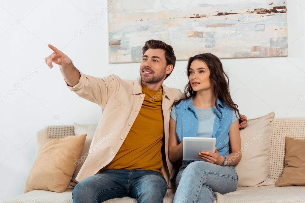Smiling man pointing with finger while woman using digital tablet in living room