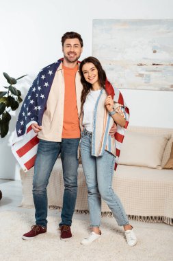 Amerikan bayrağını saran genç çift oturma odasında kameraya gülümsüyor.