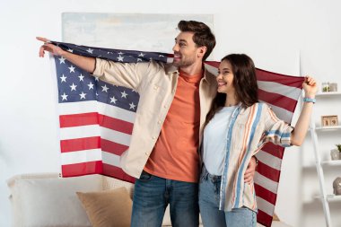 Gülümseyen adam evde Amerikan bayrağını tutarken parmak sallıyor ve kız arkadaşına sarılıyor. 