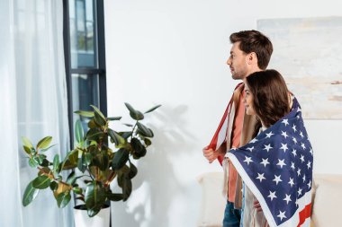Amerika bayrağına sarılmış gülümseyen bir kadın, evde erkek arkadaşının yanında duruyor.  