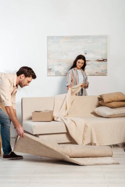 Selective focus of woman holding plaid near couch while boyfriend turning off carpet in living room clipart