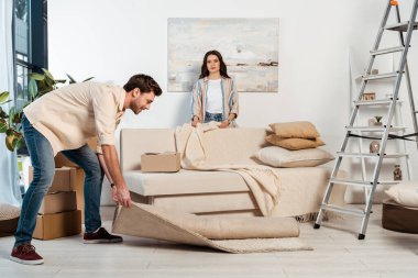 Smiling man turning off carpet near girlfriend holding plaid during relocation  clipart