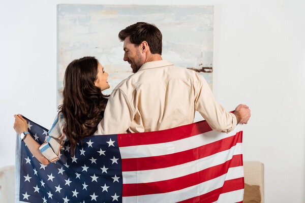 Back view of young couple smiling at each other while holding american flag at home 