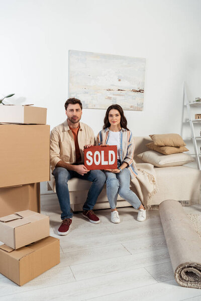 Young couple holding nameplate with sold lettering near cardboard boxes and carpet in living room