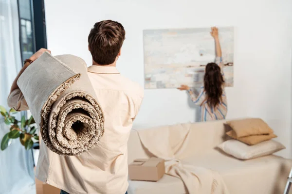 男人在搬家时拿着地毯而女朋友在墙上拿着画的背影 — 图库照片